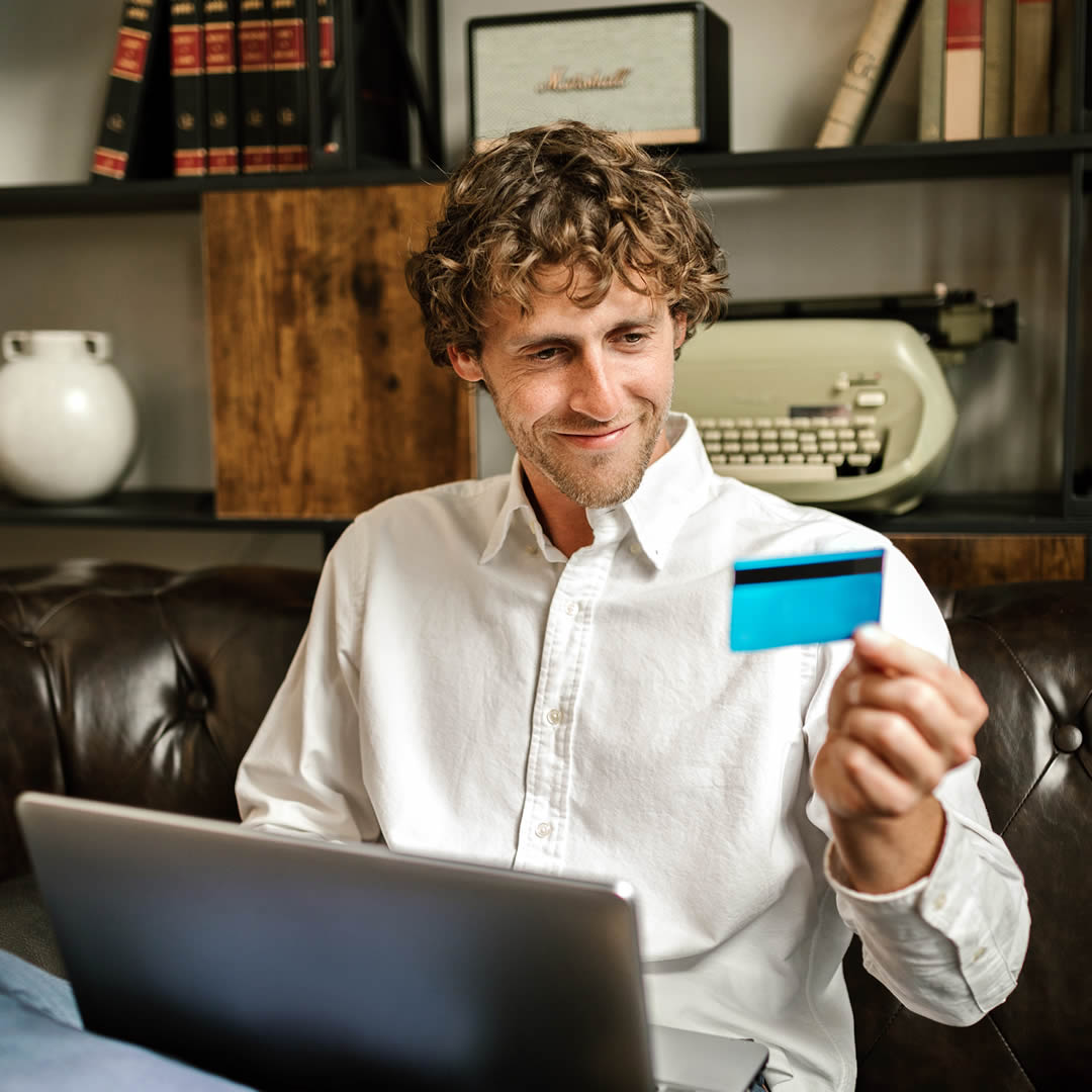 Hombre sentado con una laptop en su regazo y mirando su tarjeta de crédito