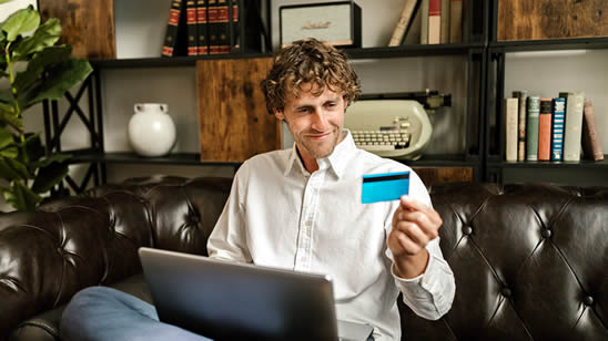 Hombre sentado con una laptop en su regazo y mirando su tarjeta de crédito